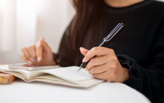 A close-up of a woman writing in a book and using her smartphone while sitting at a table indoors.
