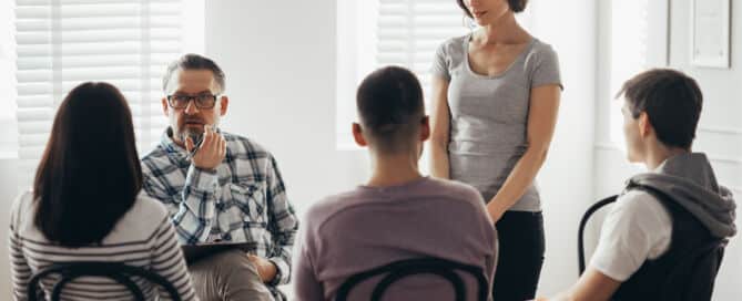 Middle age woman stands in front of a group of people during a therapy meeting and talks about her family problems