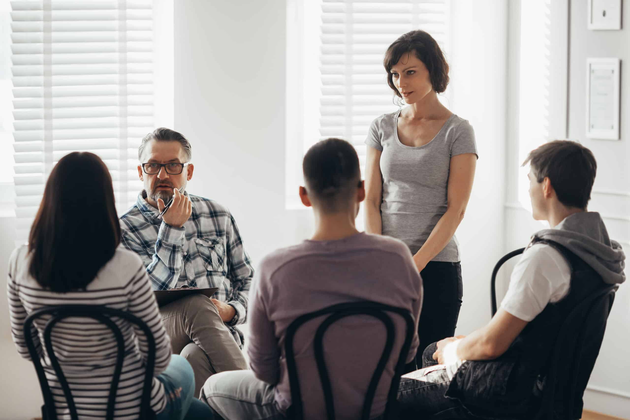 Middle age woman stands in front of a group of people during a therapy meeting and talks about her family problems